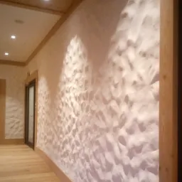 Beżowa ściana z dekoracyjną, nieregularną fakturą w korytarzu, drewniana podłoga i listwy przypodłogowe, widoczne drzwi i oświetlenie sufitowe.