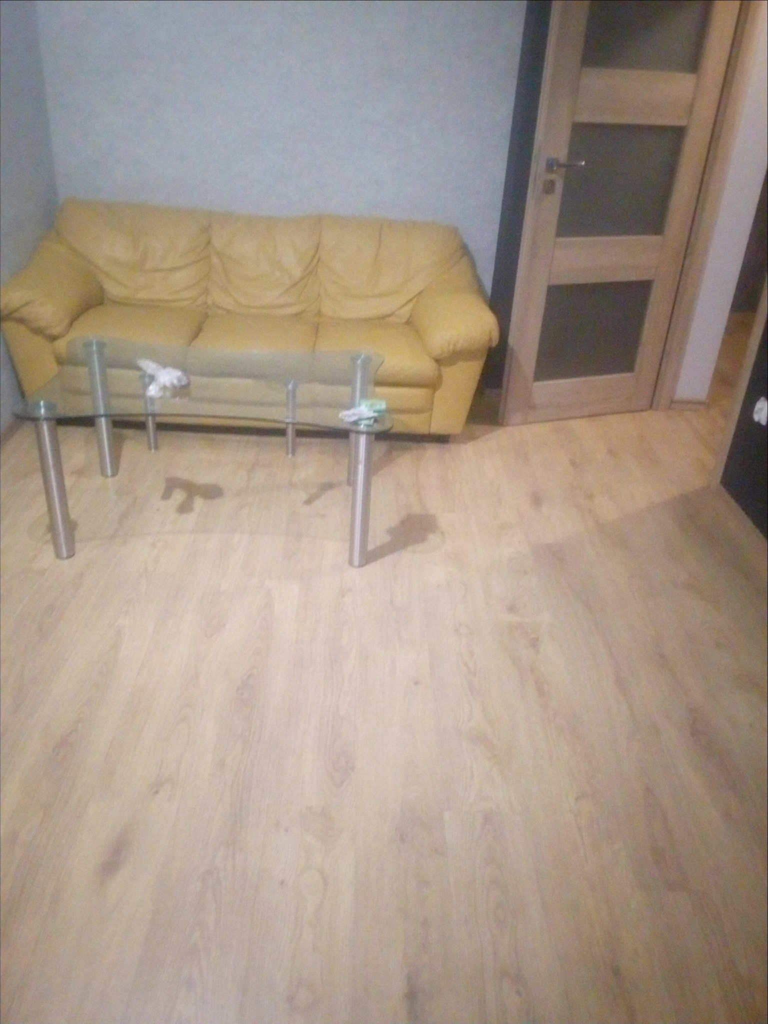 Wnętrze pokoju z żółtą, skórzaną sofą, szklanym stolikiem kawowym na metalowych nogach i jasną drewnianą podłogą, widoczne drzwi wewnętrzne z przeszkleniami.