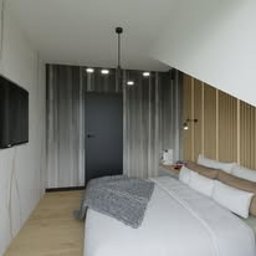 Monika Łączek IBP Custom Architecture & Design - Sypialnia na poddaszu z łóżkiem z białymi poduszkami, szarym pledem i drewnianymi panelami ściennymi, oświetlona punktowymi lampami sufitowymi i kinkietami.