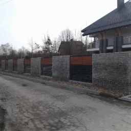Długi, kamienny mur z drewnianymi i metalowymi wstawkami oddzielający posesję od drogi, widoczny dom z szarą dachówką w tle.