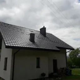 Dom jednorodzinny z nowym, ciemnym dachem z blachodachówki, dwa kominy, widoczne rynny, pochmurne niebo w tle.