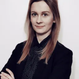 Portret uśmiechniętej prawniczki w czarnej marynarce i koszuli w kropki, z założonymi rękami na białym tle.