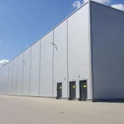 Nowoczesna hala magazynowa z szarych płyt warstwowych, widok z zewnątrz, jasne niebo z delikatnymi chmurami, widoczne bramy załadunkowe i drzwi wejściowe.