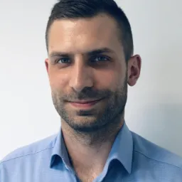 Portret mężczyzny z krótkimi, ciemnymi włosami i lekkim zarostem, ubranego w błękitną koszulę na tle białej ściany.