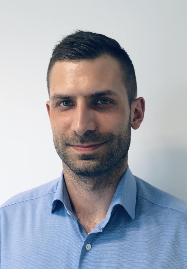 Portret mężczyzny z krótkimi, ciemnymi włosami i lekkim zarostem, ubranego w błękitną koszulę na tle białej ściany.