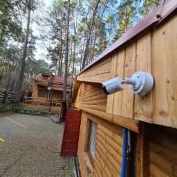 Biała kamera monitoringu zamontowana na drewnianej ścianie budynku z brązowym dachem, na tle lasu i parkingu z żółtymi liniami.