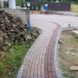 Ścieżka z kostki brukowej ułożona w łuk, obrzeża z kostki granitowej, w tle pracownik z taczką, na pierwszym planie stos kamieni i trawa.