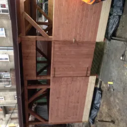 Konstrukcja drewnianej altany z widocznymi elementami zadaszenia i ścian z desek, w tle budynek mieszkalny.