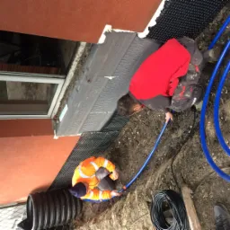 Dwóch robotników w odblaskowych ubraniach pracuje przy izolacji fundamentów budynku, układając rury drenażowe w wykopie obok ściany z oknem.