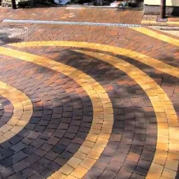 Ułożona nawierzchnia z kostki brukowej w odcieniach brązu i żółci, ułożona w koncentryczne okręgi i proste pasy, widoczna kratka ściekowa