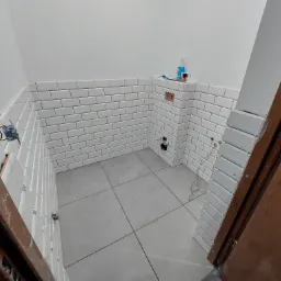 Wnętrze łazienki w trakcie remontu: białe, prostokątne płytki ułożone na ścianach do połowy wysokości, szare płytki podłogowe, widoczne narzędzia i materiały budowlane, otwór na gniazdko elektryczne.