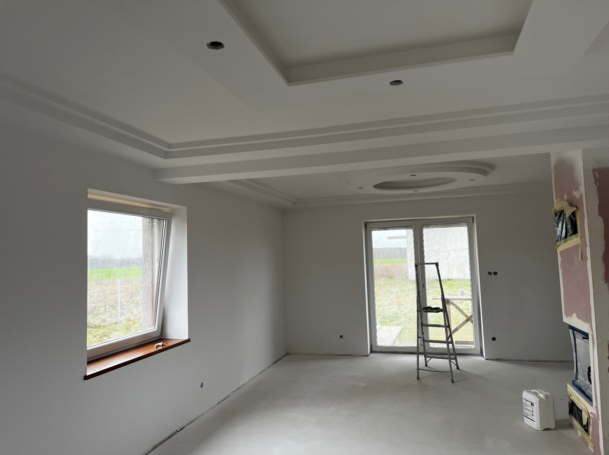 Pokój po szpachlowaniu i malowaniu na biało, widoczna drabina przy oknie balkonowym, gładka powierzchnia ścian i sufitu z dekoracyjnymi elementami, otwory na oświetlenie.