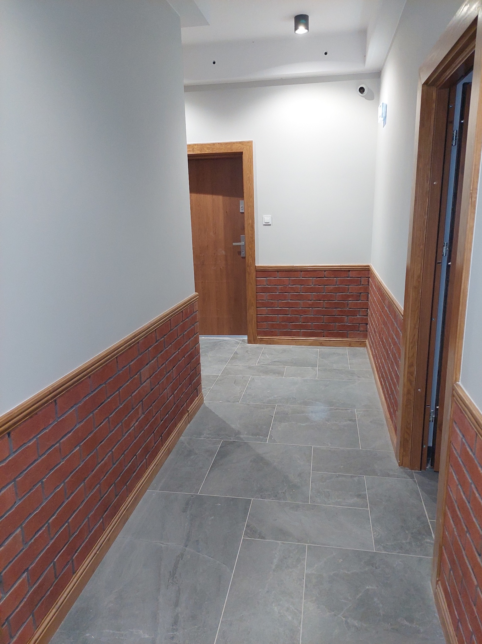 Korytarz z szarymi płytkami podłogowymi, ściany pomalowane na szaro z dekoracyjną cegłą do połowy wysokości, drewniane futryny drzwi.