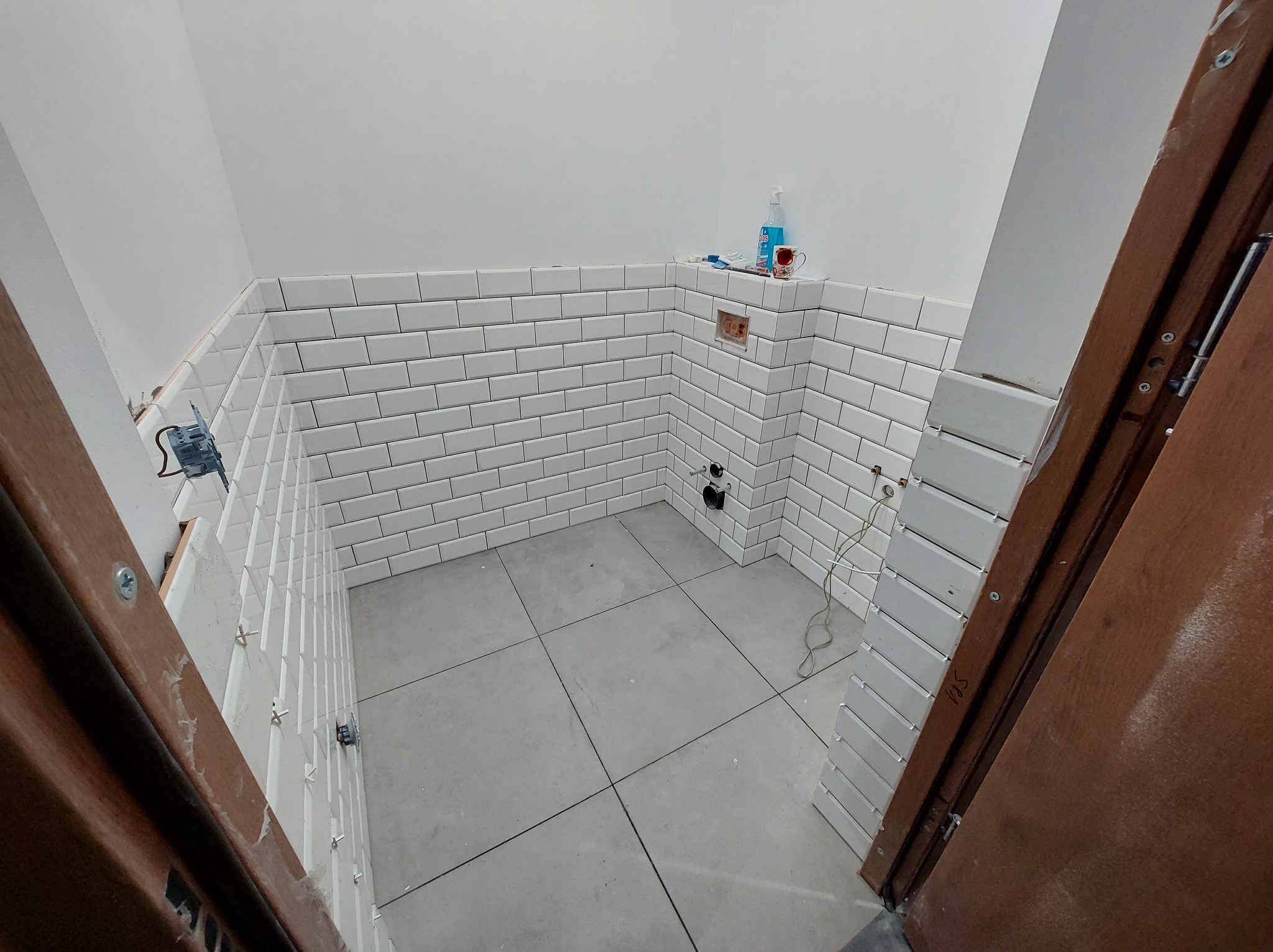 Wnętrze łazienki w trakcie remontu: białe, prostokątne płytki ułożone na ścianach do połowy wysokości, szare płytki podłogowe, widoczne narzędzia i materiały budowlane, otwór na gniazdko elektryczne.