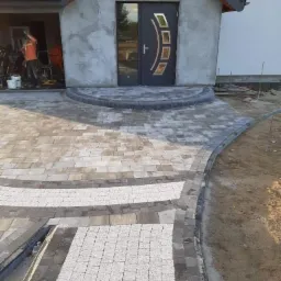 Ułożona kostka brukowa przed wejściem do domu z ciemnymi drzwiami. Szare i białe kostki tworzą wzór. Widoczne elementy wykończeniowe i narzędzia w tle.