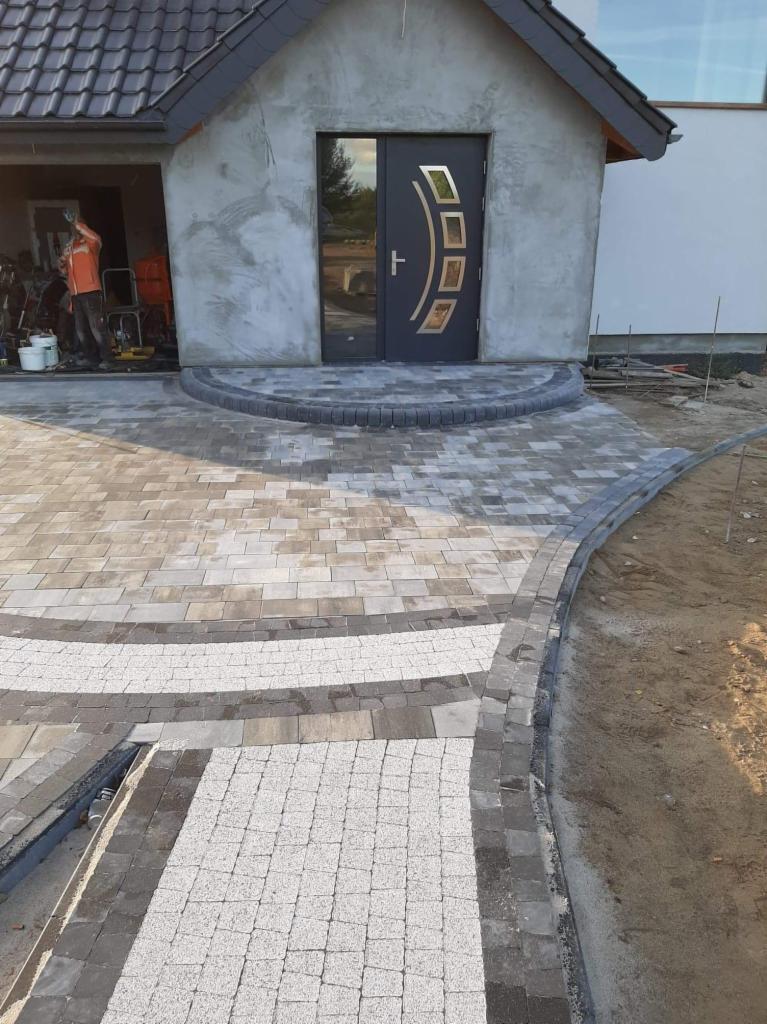Ułożona kostka brukowa przed wejściem do domu z ciemnymi drzwiami. Szare i białe kostki tworzą wzór. Widoczne elementy wykończeniowe i narzędzia w tle.