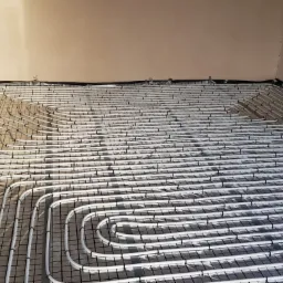 Białe rury ogrzewania podłogowego, ułożone w pętle na czarnej siatce, przed wylaniem posadzki, widoczne elementy mocujące przy ścianie.