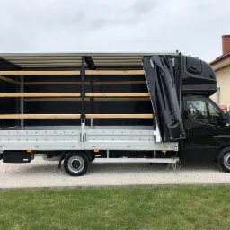 Czarny bus z odkrytą plandeką boczną, widoczne drewniane poprzeczki wewnątrz przestrzeni ładunkowej, zaparkowany na żwirowej drodze przed domem.