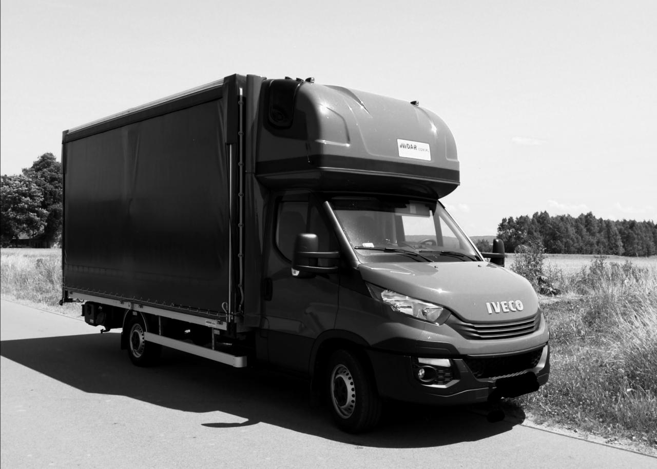 Czarno-białe zdjęcie samochodu dostawczego Iveco z plandeką na tle polnej drogi i drzew, widoczne logo 'WIDAR'.