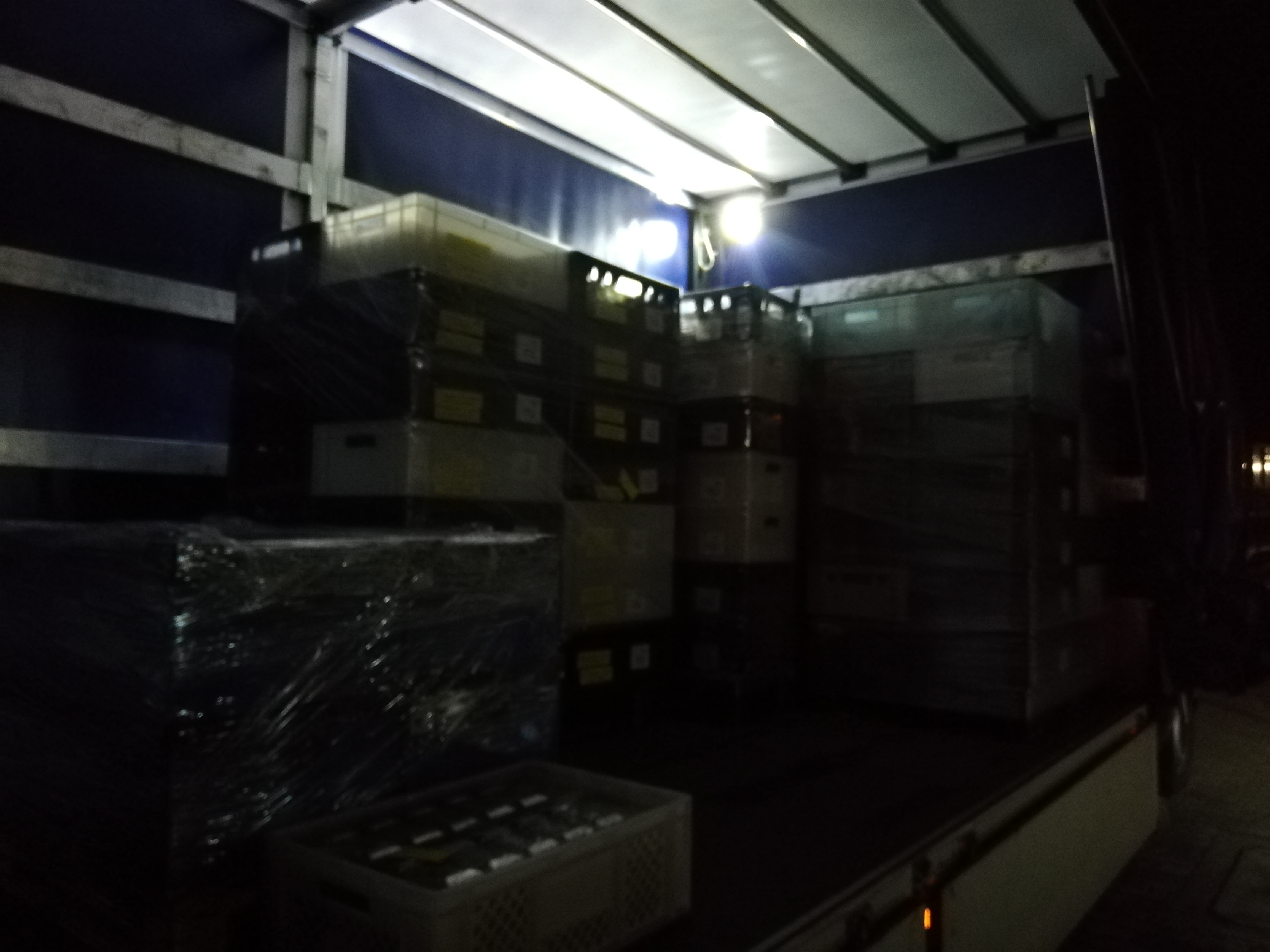 Wnętrze ciężarówki wypełnionej kartonami i skrzynkami, niektóre owinięte folią, oświetlone słabym światłem sufitowym.