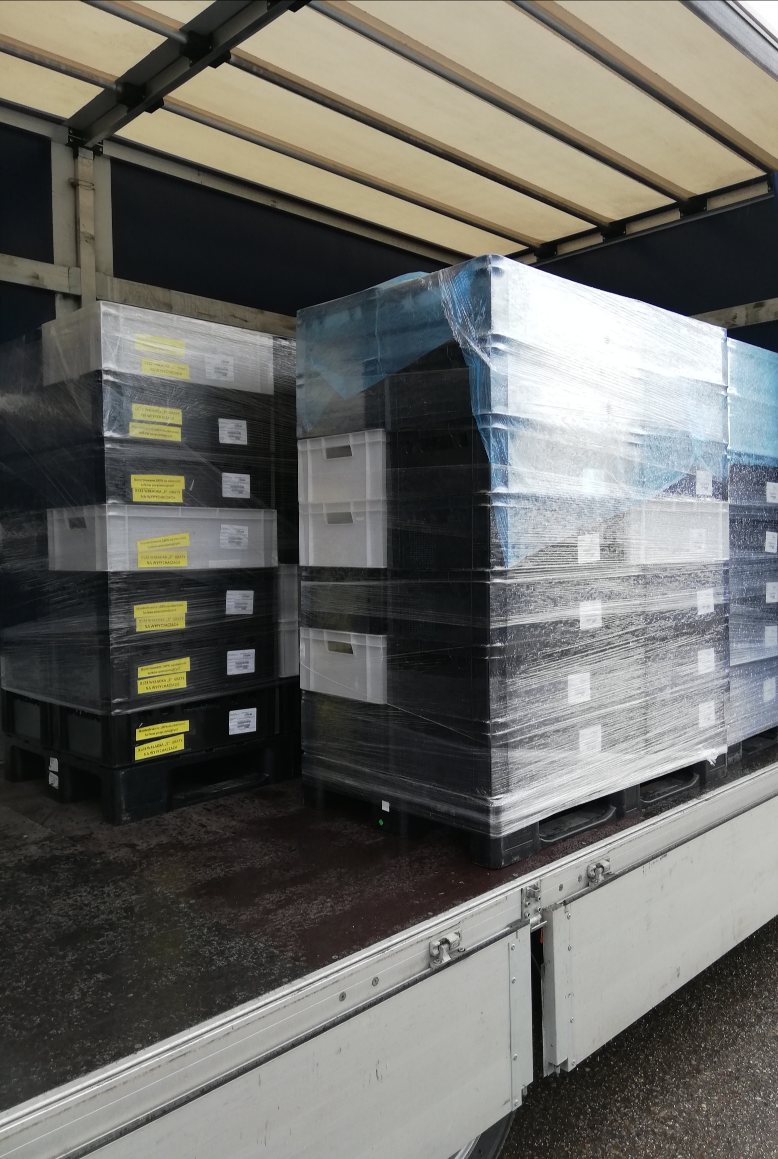 Wnętrze naczepy z załadowanymi paletami czarno-białych plastikowych pojemników, zabezpieczonych folią stretch, widoczne etykiety na pojemnikach.