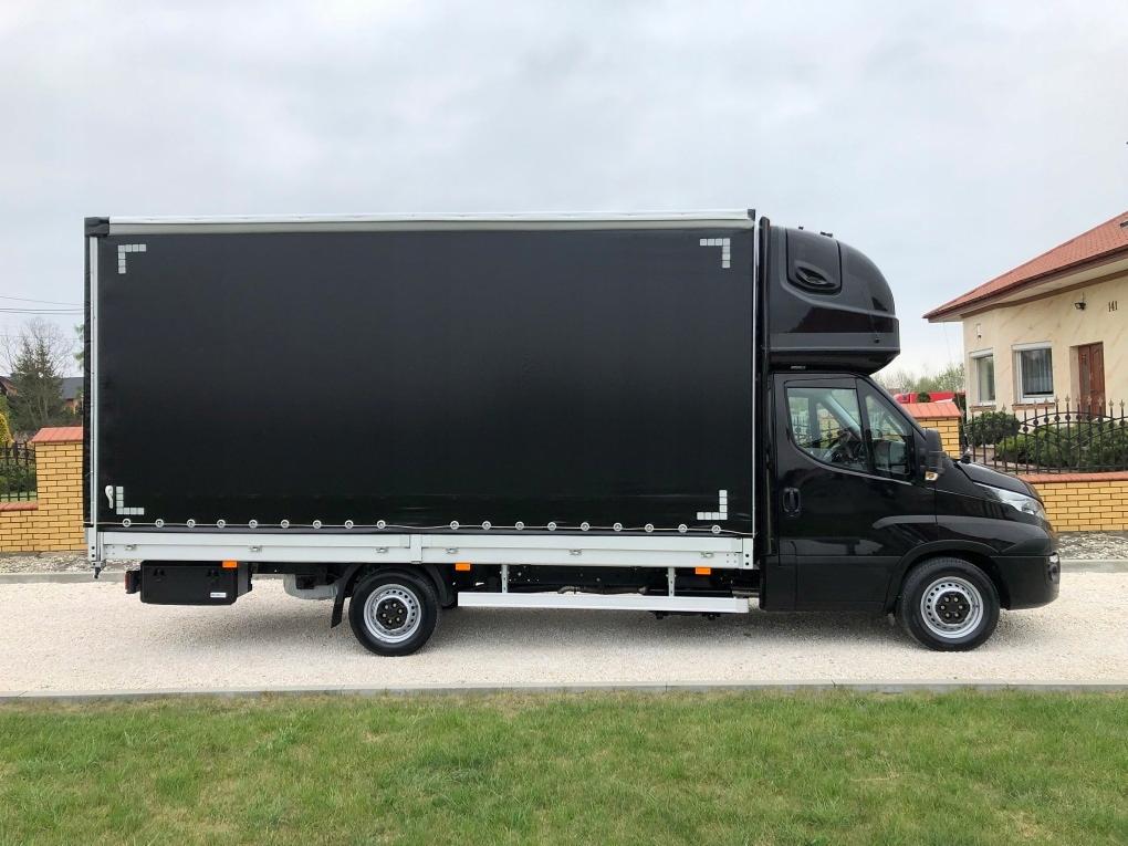 Czarny samochód dostawczy z plandeką, zaparkowany na kostce brukowej przed domem z ceglanym płotem, widok z boku.