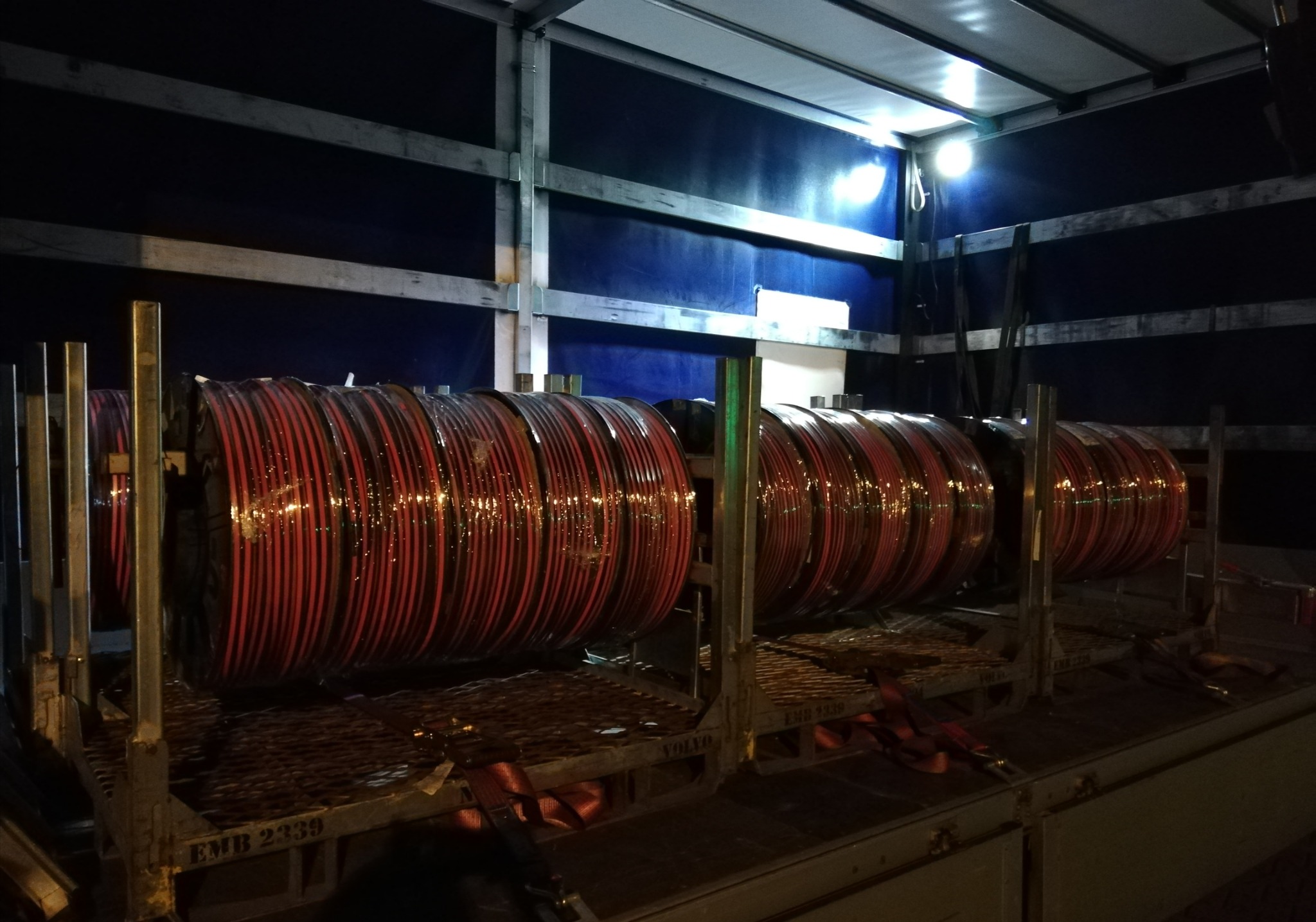 Zabezpieczone metalowe szpule z czerwonymi kablami wewnątrz naczepy ciężarówki, oświetlone górnym światłem.