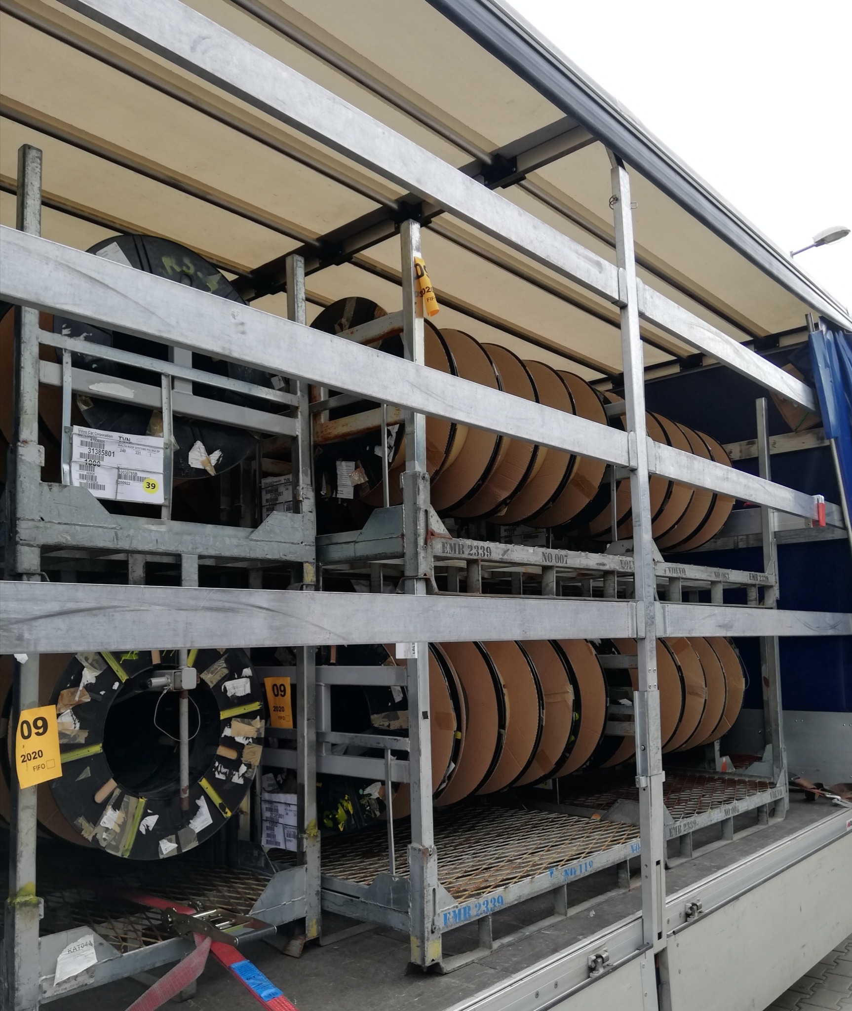 Wnętrze naczepy z metalowymi regałami, na których umieszczone są rolki z materiałem, zabezpieczone etykietami i taśmami, widoczne oznaczenia 'EMB 2339' i 'Volvo Car Corporation'.
