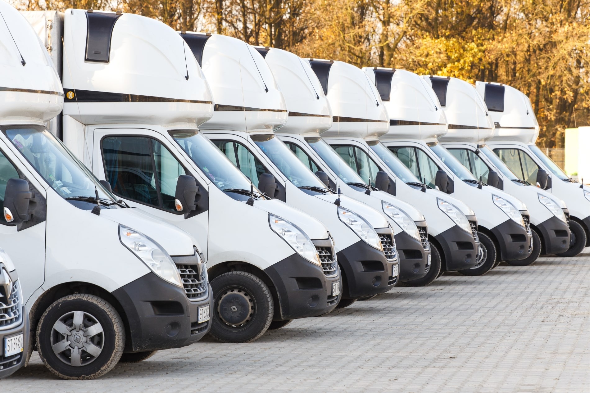 Rząd identycznych białych busów z nadbudówkami, zaparkowanych na brukowanym placu, z jesiennymi drzewami w tle.