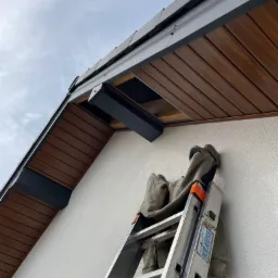 Montaż podbitki dachowej z drewnianymi panelami i metalowymi elementami wykończeniowymi, widoczna drabina oparta o ścianę budynku.