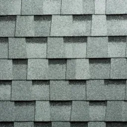 Szczegółowe ujęcie szarej, teksturowanej powierzchni dachu pokrytej bitumicznymi płytkami w kształcie prostokątów, układających się w regularny wzór z widocznymi cieniami.