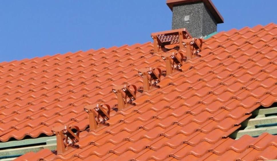 Pomarańczowy dach z dachówki ceramicznej, zabezpieczony metalowymi uchwytami przeciwśniegowymi, z widocznym kominem na tle błękitnego nieba.