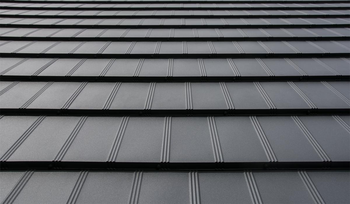 Szare, metalowe pokrycie dachowe ułożone w rzędach, z wyraźnymi pionowymi przetłoczeniami i czarnymi listwami łączeniowymi.