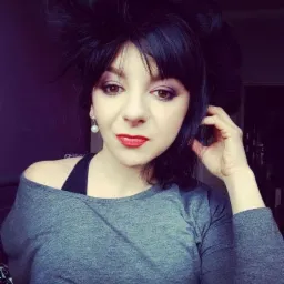 Portret kobiety z ciemnymi włosami, wyrazistym makijażem i perłową kolczykiem. Uczesanie z uniesionymi włosami dodaje charakteru. Ciemne tło.