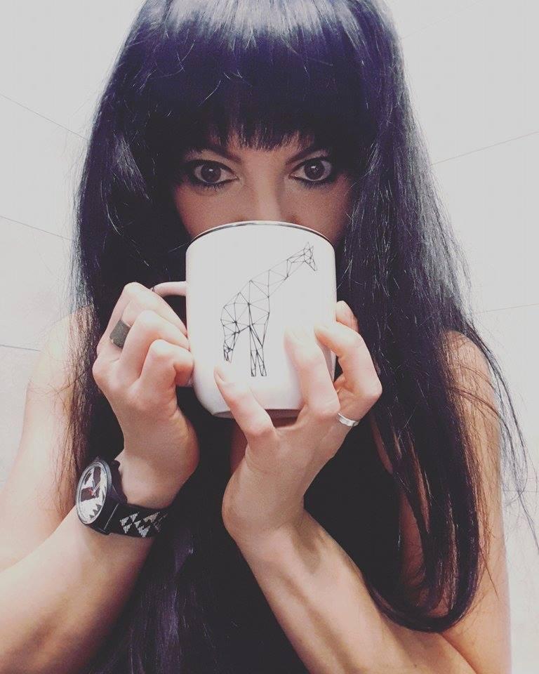 Kobieta z czarnymi włosami pije z białego kubka z geometryczną żyrafą, widoczny zegarek i pierścionki.