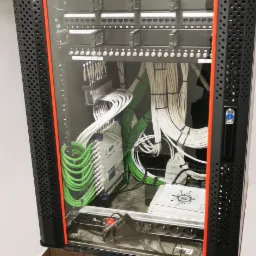 Kompletna szafa rack z precyzyjnie ułożonymi kablami w kolorach białym i zielonym, widoczne moduły i złącza, całość zamknięta w perforowanej obudowie z pomarańczowymi akcentami.