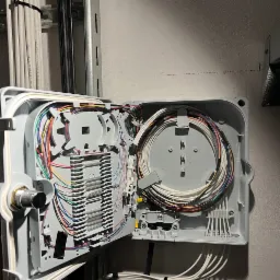 Otwarta skrzynka z kablami światłowodowymi na ścianie, uporządkowane przewody wewnątrz szarej obudowy, widoczne złącza i elementy montażowe.