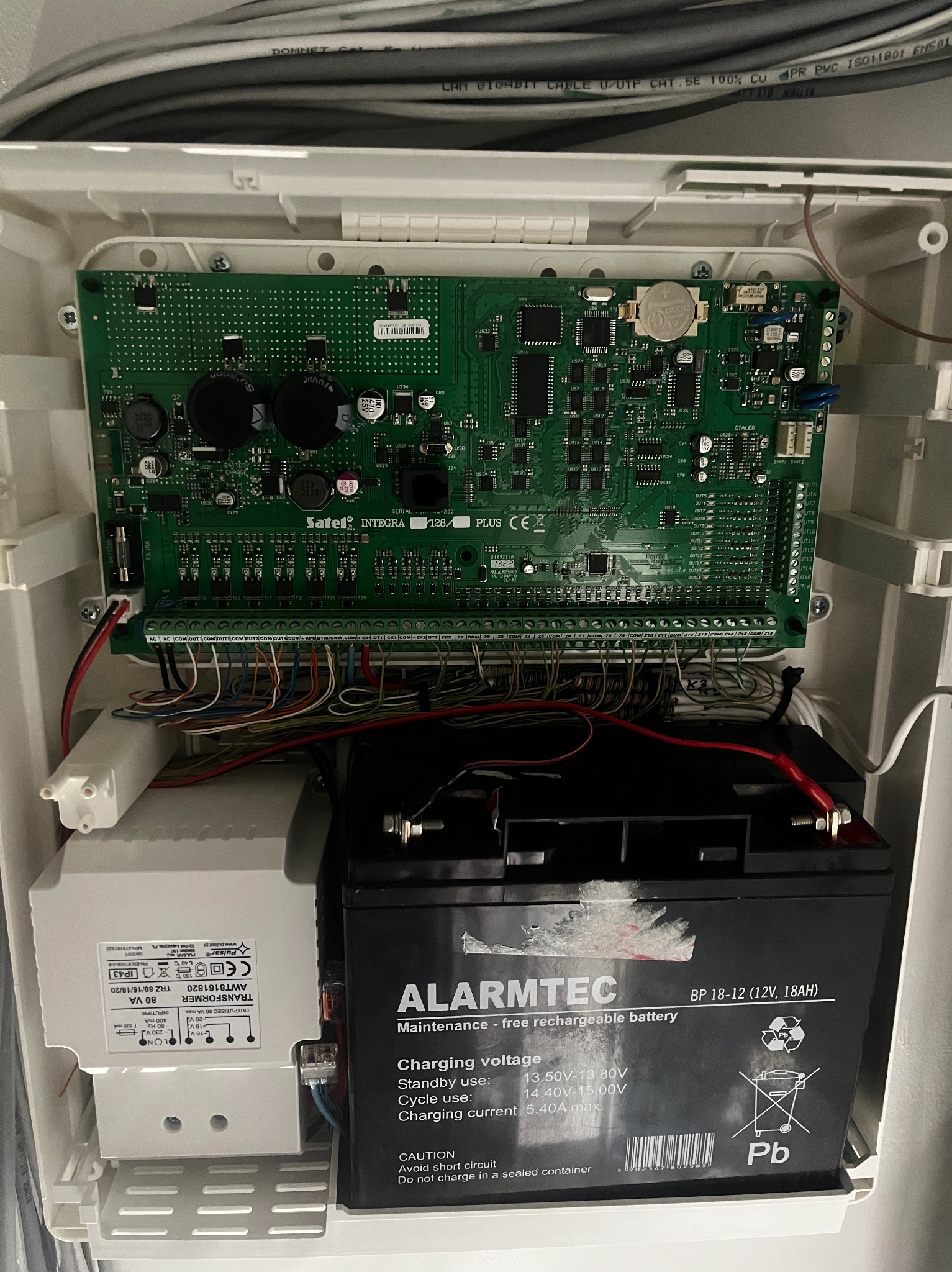 Centrala alarmowa Satel Integra 128 Plus z akumulatorem Alarmtec BP 18-12 w białej obudowie. Widoczne kable i transformator Pulsar AWT8161820.