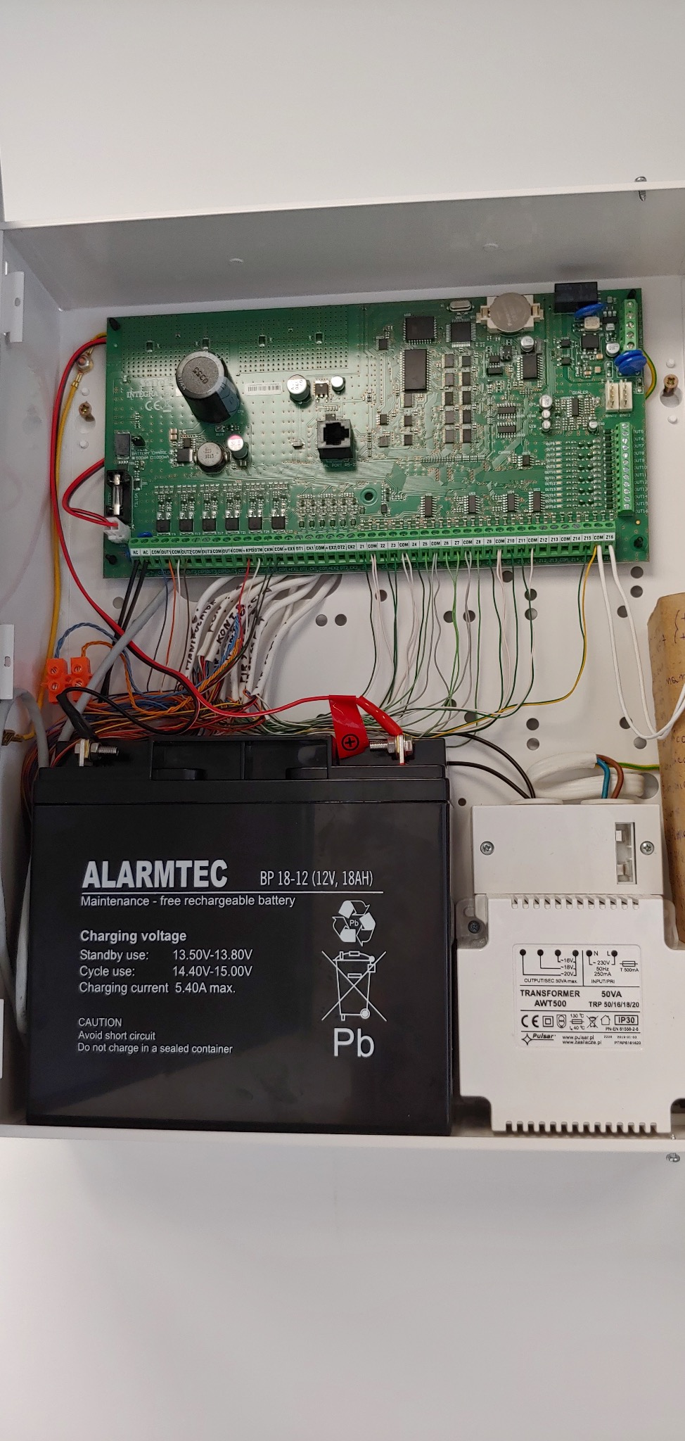 Szafka z elektroniką alarmową: akumulator Alarmtec, transformator Pulsar AWT500 i zielona płyta główna z licznymi przewodami. Widok na wnętrze szafki.