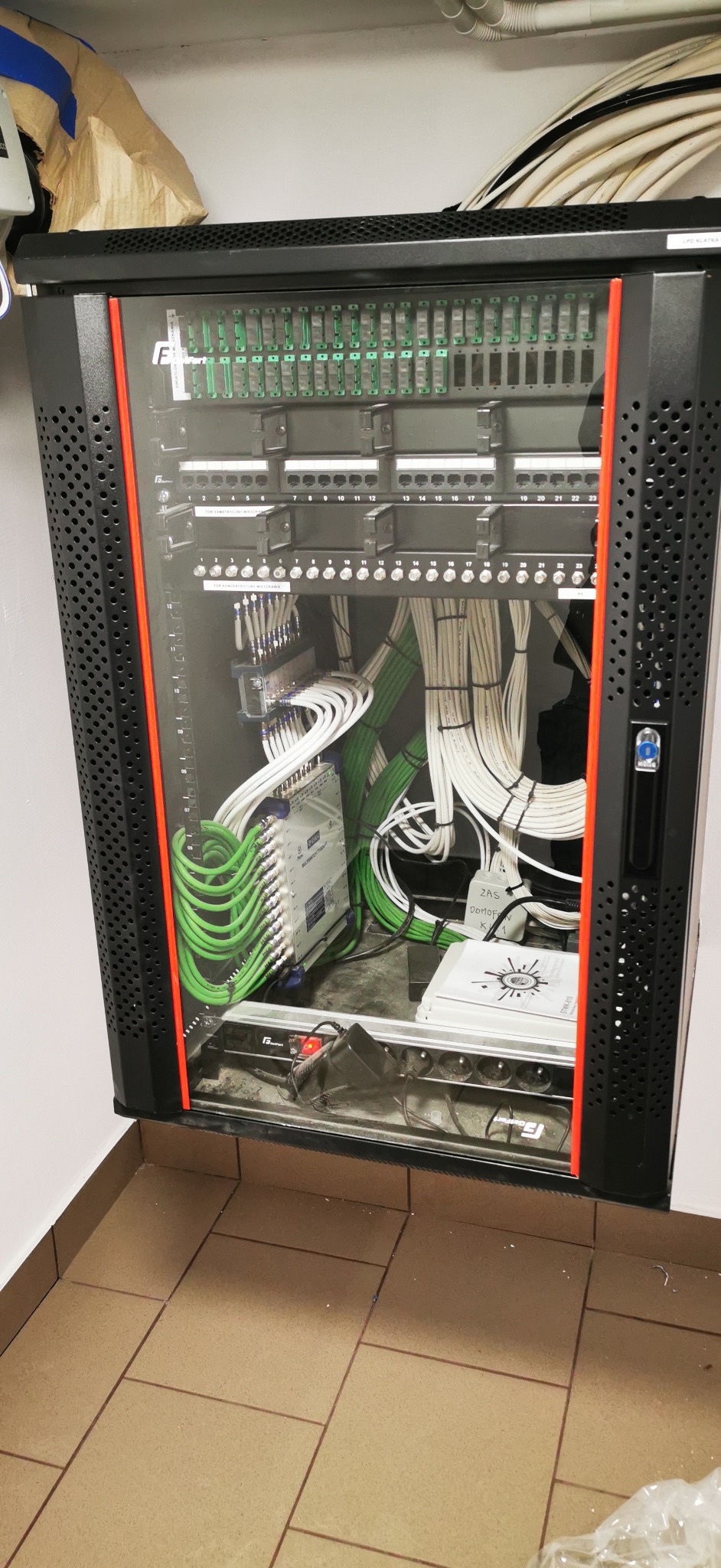 Kompletna szafa rack z precyzyjnie ułożonymi kablami w kolorach białym i zielonym, widoczne moduły i złącza, całość zamknięta w perforowanej obudowie z pomarańczowymi akcentami.
