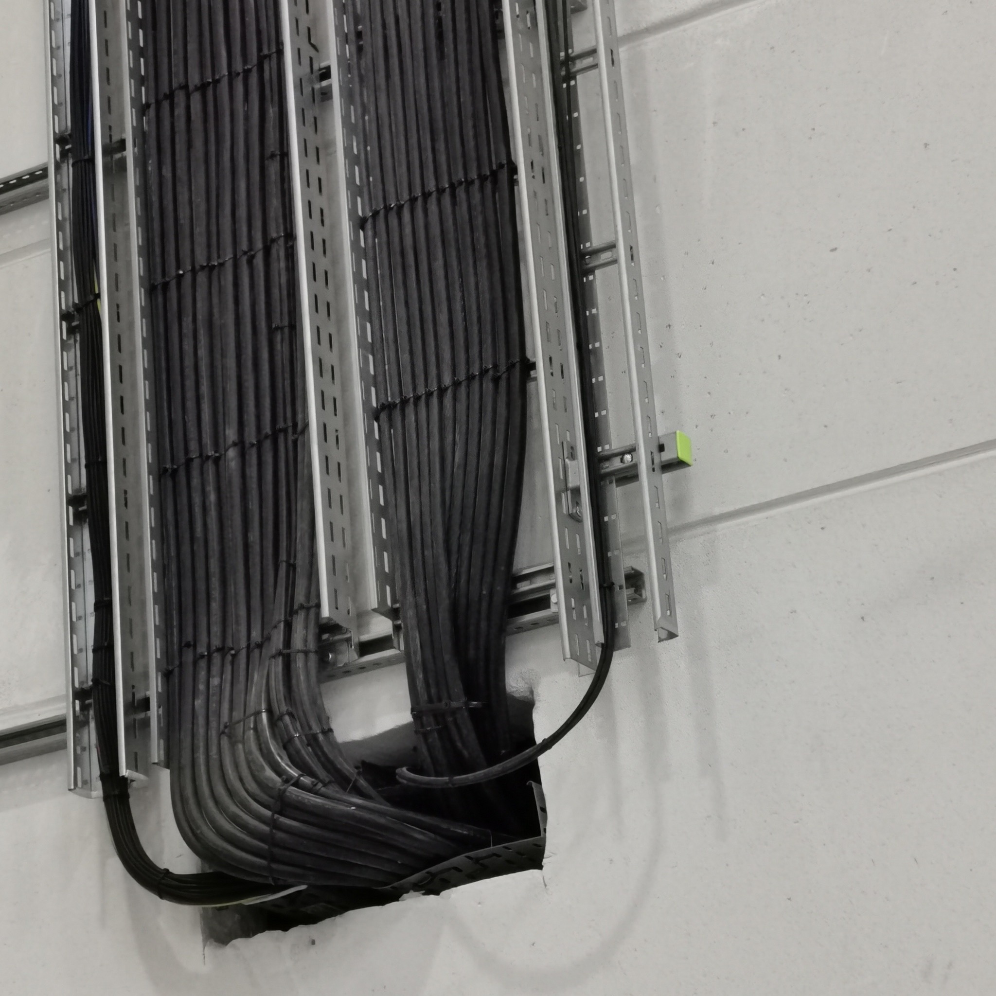 Rząd kabli w metalowych korytkach, wchodzących w otwór w ścianie. Szare tło ściany. Kable ułożone równolegle. Widok z bliska na instalację elektryczną.