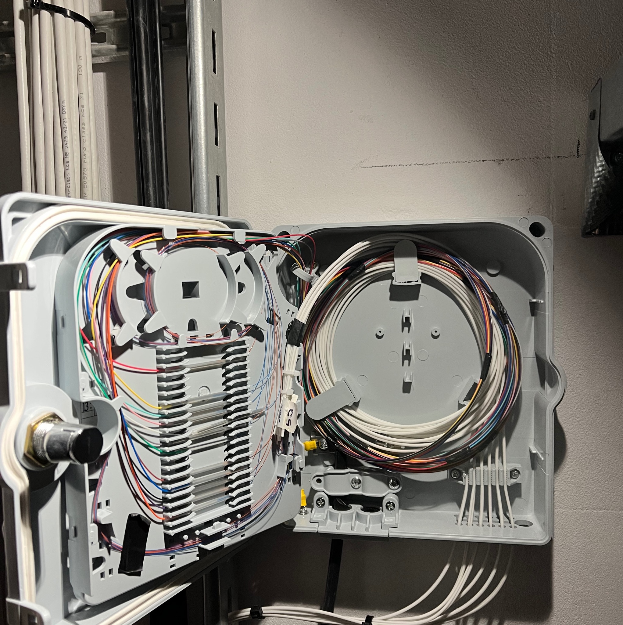 Otwarta skrzynka z kablami światłowodowymi na ścianie, uporządkowane przewody wewnątrz szarej obudowy, widoczne złącza i elementy montażowe.