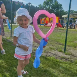 Dziewczynka w białym kapeluszu trzyma balon w kształcie serca i niebieski balon skręcony w spiralę, w tle plac zabaw i inne dzieci.