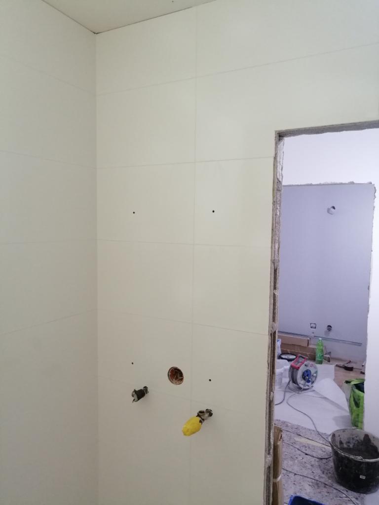 Ściana w łazience wyłożona białymi płytami gipsowo-kartonowymi, z widocznymi liniami podziału, przygotowana pod okładzinę, z wystającymi przyłączami wodnymi i elektrycznymi oraz otworem, widok...