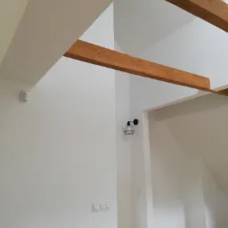 Wnętrze po remoncie z widocznymi drewnianymi belkami stropowymi, białą ścianą z zamontowanym czujnikiem ruchu i włącznikami światła oraz nowoczesną lampą ścienną, częściowo zakryte rolką papieru...