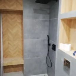 Nowoczesna łazienka z szarymi płytkami, deszczownicą w kolorze czarnym, wnęką wykończoną drewnem w jodełkę i ukrytym systemem spłukiwania