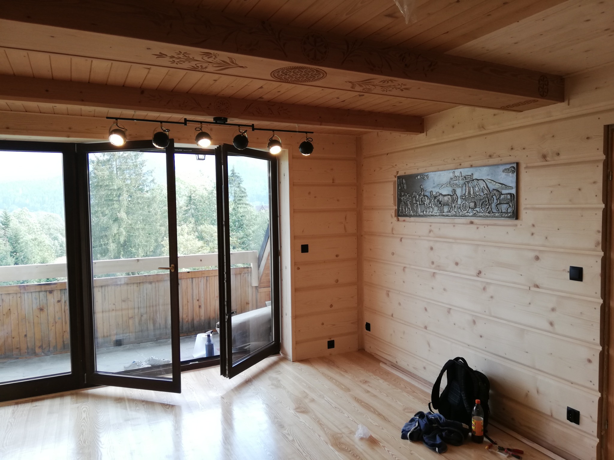 Wnętrze pokoju wykończonego boazerią z jasnego drewna, z otwartymi drzwiami balkonowymi, widokiem na zalesione wzgórza, dekoracyjnym wzorem na belkach sufitowych i metalową płaskorzeźbą na ścianie.