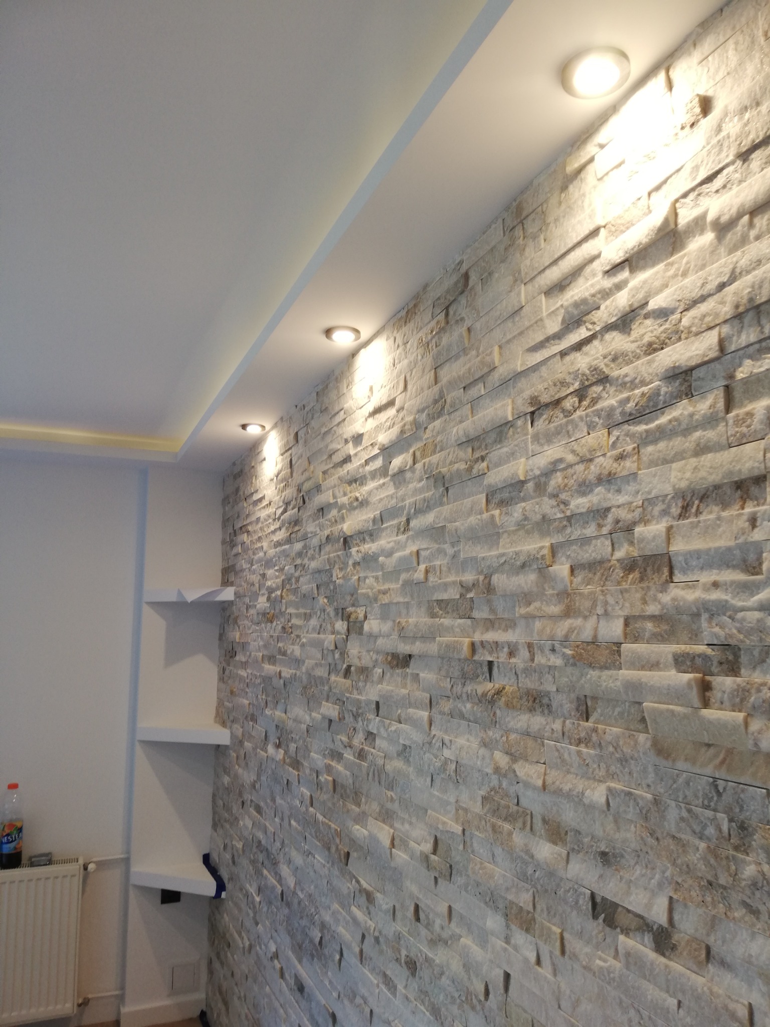 Ściana wyłożona dekoracyjnym kamieniem w odcieniach szarości i beżu, oświetlona trzema punktowymi lampami halogenowymi w suficie podwieszanym, z elementami zabudowy półek po lewej stronie.