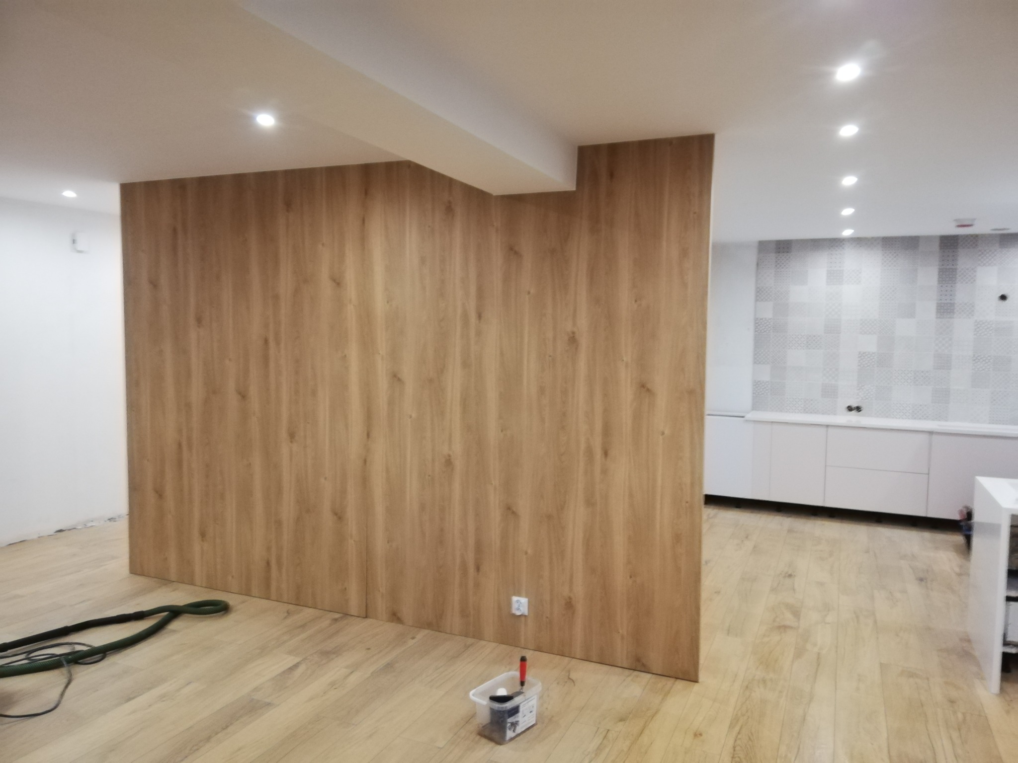 Wnętrze w trakcie prac wykończeniowych: ściana obłożona panelami drewnopodobnymi, widoczna instalacja elektryczna, podłoga z paneli, w tle zabudowa kuchenna z mozaiką na ścianie, narzędzia...