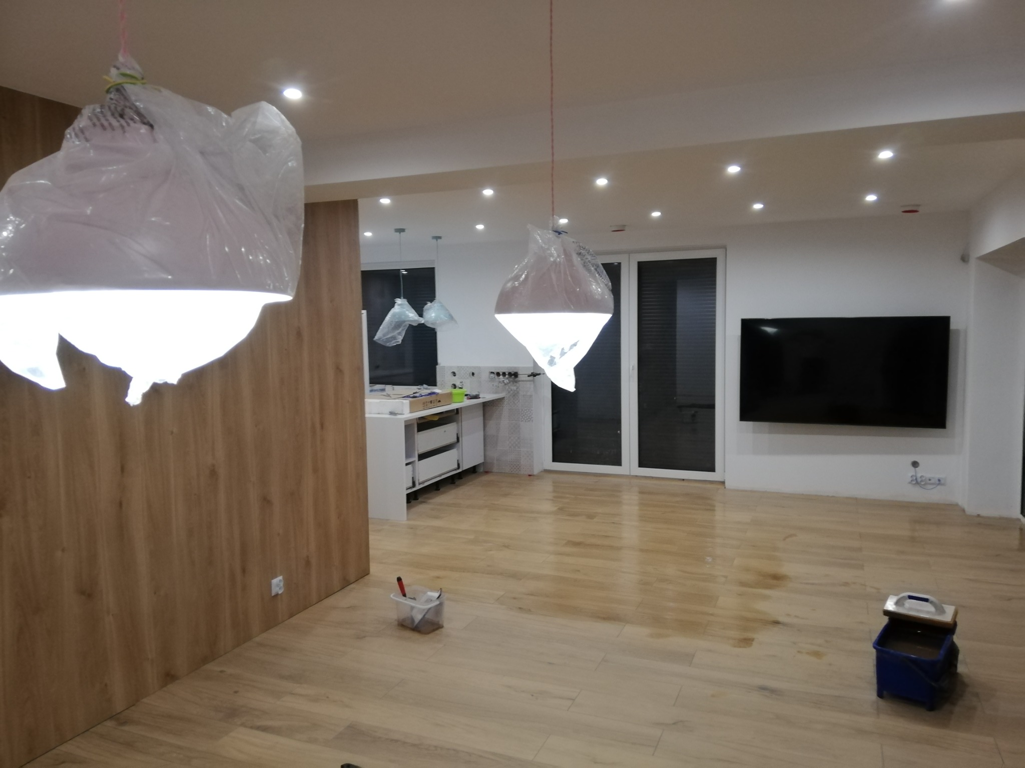 Wnętrze w trakcie prac wykończeniowych: podłoga wyłożona panelami imitującymi drewno, lampy zabezpieczone folią, ślady po pracach na podłodze, widoczne narzędzia malarskie i wiadro.
